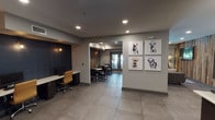 Located at 2121 Delgany St in Denver's vibrant LoDo neighborhood, this single-floor amenity space at The Metro Urban Apartments is a thoughtfully designed communal retreat. Guests are welcomed through glass-paneled entry doors bearing the property's logo, setting a polished first impression. Large-format gray tile flooring flows throughout, anchoring the open-plan layout that seamlessly connects multiple functional zones. The lounge area is a standout feature, centered around a reclaimed wood accent wall that adds warmth and texture, complemented by a wall-mounted flat-screen TV, tan leather armchairs, and a rustic wood coffee table. Floor-to-ceiling windows flood this seating area with natural light while offering views of outdoor greenery. Adjacent to the lounge, a dining and workspace area provides bistro-style tables and a built-in workstation with a desktop computer, ideal for residents needing a quiet spot to focus. The fully equipped kitchen features white subway tile backsplash, dark cabinetry, white quartz countertops, open shelving, stainless steel appliances, and a reclaimed wood accent wall echoing the lounge's aesthetic. Playful bulldog artwork adorns the walls, injecting personality into the space. Recessed lighting throughout ensures a warm, welcoming ambiance across all areas.