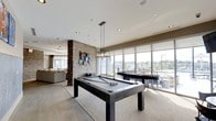 This amenity floor offers a thoughtfully designed social and recreational space that combines modern aesthetics with functional comfort. The game room serves as a central highlight, featuring a full-size billiards table illuminated by an industrial-style pendant light fixture, creating a focal point that draws residents in. A second gaming table nearby adds to the entertainment options available. Expansive floor-to-ceiling windows line the perimeter, flooding the space with abundant natural light and offering sweeping views of the surrounding cityscape and outdoor terrace, where patio seating is visible. The open-concept layout seamlessly connects the game area to a comfortable lounge zone, furnished with plush sofas and defined by an exposed brick accent column adorned with decorative wall art, adding texture and character to the interior. Recessed lighting throughout the ceiling complements the natural light, ensuring a bright and welcoming atmosphere at any hour. Neutral tones in the flooring, furnishings, and walls create a cohesive, contemporary aesthetic, while roller shades on the windows offer adjustable light control. The overall ambiance is modern, inviting, and well-suited for both social gatherings and casual relaxation.