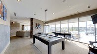 This amenity floor offers a thoughtfully designed social and recreational space that combines modern aesthetics with functional comfort. The game room serves as a central highlight, featuring a full-size billiards table illuminated by an industrial-style pendant light fixture, creating a focal point that draws residents in. A second gaming table nearby adds to the entertainment options available. Expansive floor-to-ceiling windows line the perimeter, flooding the space with abundant natural light and offering sweeping views of the surrounding cityscape and outdoor terrace, where patio seating is visible. The open-concept layout seamlessly connects the game area to a comfortable lounge zone, furnished with plush sofas and defined by an exposed brick accent column adorned with decorative wall art, adding texture and character to the interior. Recessed lighting throughout the ceiling complements the natural light, ensuring a bright and welcoming atmosphere at any hour. Neutral tones in the flooring, furnishings, and walls create a cohesive, contemporary aesthetic, while roller shades on the windows offer adjustable light control. The overall ambiance is modern, inviting, and well-suited for both social gatherings and casual relaxation.