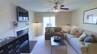 This single-floor space centers around a well-appointed living room that balances comfort and functionality. The room is anchored by a sleek fireplace with a dark stone surround, complemented by a built-in shelf above that provides display space. Neutral, warm-toned walls create a calm and inviting atmosphere throughout.

A ceiling fan with an integrated light fixture is centrally positioned overhead, ensuring both air circulation and ambient lighting. The carpeted floor adds warmth and softness underfoot, contributing to the room's relaxed character. A large window along the back wall allows natural light to filter into the space, brightening the interior and providing a view of the outdoors.

The living area is furnished with a pair of plush sofas accented with decorative pillows in green and neutral tones, arranged around a dark rectangular coffee table that serves as a practical centerpiece. Floor lamps on either side of the room provide additional soft lighting. The overall layout is thoughtfully arranged to encourage conversation and relaxation, making this a welcoming and functional living environment.
