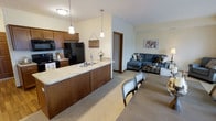 This single-floor residence offers an open-concept layout that seamlessly connects the kitchen, dining, and living areas into one cohesive and functional space. The kitchen is well-appointed with rich, warm-toned wood cabinetry, a light granite countertop, and black appliances including a refrigerator, range, and microwave. A kitchen peninsula with an undermount sink serves as both a practical workspace and a natural divider, while two pendant lights hang above to provide focused task lighting and add a stylish touch. Adjacent to the kitchen, the dining area accommodates a full table and chairs, making it ideal for everyday meals or casual gatherings. The living room extends naturally from the dining space, featuring neutral carpet flooring and soft, light-colored walls that create a bright and airy atmosphere. The space is finished with warm wood baseboards throughout, adding a consistent design accent. Overall, the open floor plan fosters a sense of spaciousness and ease of movement, while the neutral palette and warm wood tones work together to create an inviting and comfortable environment suited for everyday living.