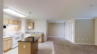 This single-floor residence showcases an open-concept layout that seamlessly connects the kitchen and living areas, creating a cohesive and functional living environment. The kitchen is outfitted with light maple cabinetry, granite-style countertops, and a double basin sink positioned beneath upper cabinets. A pendant light fixture hangs above, providing focused task lighting, while a dark-surfaced kitchen island or breakfast bar adds a contrasting design element and additional workspace in the foreground.

The flooring transitions from tile in the kitchen area to plush neutral-toned carpet throughout the living space, adding warmth and comfort underfoot. The living area is open and generously proportioned, with neutral beige walls and standard-height ceilings that contribute to a clean, understated aesthetic. A set of vertical blinds covers what appears to be a sliding glass door, suggesting access to an outdoor area and a source of natural light. Recessed lighting in the living area supplements the overall illumination. The uncluttered, move-in-ready condition of the space allows its layout and features to be clearly appreciated.