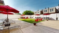 Located at 2746 Belmont Ave in Philadelphia's Wynnefield neighborhood, this outdoor amenity space is thoughtfully designed for relaxation and social gathering. The grounds feature a well-maintained central lawn area framed by wide concrete patio surfaces that extend along the perimeter, offering ample room for both active use and quiet retreat. Vibrant red Adirondack chairs are positioned throughout the grass, providing casual seating that complements the lively atmosphere. Multiple dining sets, each shaded by bold red market umbrellas, are arranged along the patio edges, ideal for outdoor meals or conversation. Chaise lounges offer additional options for sun-soaking or unwinding. A dedicated lounge seating area with navy blue sofas and accent chairs sits against the warm tan brick exterior of the adjacent multi-story building, creating a more intimate courtyard feel. The entire space is enclosed by a tall white privacy fence, ensuring a secure and peaceful environment. Potted plants and greenery are scattered throughout, adding natural color and texture. Grills are available for outdoor cooking, enhancing the functionality of the space. Open to blue skies and natural sunlight, this outdoor area serves as a versatile communal retreat within an urban residential setting.