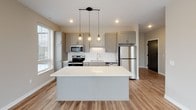 Located at 229 Roberts St N in downtown Fargo, North Dakota, this single-floor unit offers a thoughtfully designed open-concept layout with contemporary finishes throughout. The kitchen serves as the centerpiece of the living space, showcasing gray shaker-style cabinetry, white quartz countertops, a herringbone tile backsplash, and a full suite of stainless steel appliances including an electric range, microwave, dishwasher, and refrigerator. A large central island with pendant cage lighting above provides additional prep space and a natural gathering point that flows seamlessly into the main living area. Warm wood-look flooring runs throughout the main living and kitchen spaces, adding a welcoming contrast to the crisp white finishes. The bedroom features plush carpeting and large windows that frame views of the surrounding downtown Fargo skyline, filling the room with natural light. The bathroom is appointed with elegant marble-style tile flooring, a vanity with a white countertop, and black hardware accents that add a modern touch. A dedicated laundry area houses a stacked full-size washer and dryer unit alongside built-in wire shelving for added storage convenience. The overall aesthetic is clean, modern, and functional, making this a well-appointed urban residence in the heart of Fargo.
