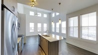 This single-floor residence at Overture showcases a thoughtfully designed open-concept layout that seamlessly connects the entryway, kitchen, and living areas. The space is immediately distinguished by its dramatically high ceilings, which rise well above standard height and are lined with a row of clerestory windows that flood the interior with natural light from multiple angles. Large windows along the perimeter walls further enhance the bright, airy atmosphere.

The kitchen serves as the focal point of the main living area, anchored by a generously sized island featuring a double sink, stone countertops, and warm wood cabinetry. Stylish pendant lights hang above the island, complementing a ceiling fan and additional track lighting overhead. Stainless steel appliances complete the modern kitchen aesthetic.

Dark hardwood-style flooring runs throughout the space, providing a rich contrast to the crisp white walls and bright ceiling. A louvered door near the entry suggests convenient storage or utility access. The open sightlines from the entry corridor to the living area create a welcoming sense of depth and spaciousness, making this a bright and functional modern residence.