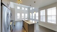 This single-floor residence at Overture showcases a thoughtfully designed open-concept layout that seamlessly connects the entryway, kitchen, and living areas. The space is immediately distinguished by its dramatically high ceilings, which rise well above standard height and are lined with a row of clerestory windows that flood the interior with natural light from multiple angles. Large windows along the perimeter walls further enhance the bright, airy atmosphere.

The kitchen serves as the focal point of the main living area, anchored by a generously sized island featuring a double sink, stone countertops, and warm wood cabinetry. Stylish pendant lights hang above the island, complementing a ceiling fan and additional track lighting overhead. Stainless steel appliances complete the modern kitchen aesthetic.

Dark hardwood-style flooring runs throughout the space, providing a rich contrast to the crisp white walls and bright ceiling. A louvered door near the entry suggests convenient storage or utility access. The open sightlines from the entry corridor to the living area create a welcoming sense of depth and spaciousness, making this a bright and functional modern residence.