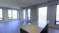 This single-floor unit is situated at a significant elevation within a high-rise building, offering commanding panoramic views of the surrounding cityscape through its expansive, floor-to-ceiling windows. The open-concept layout seamlessly connects the kitchen and living areas, creating a spacious and airy atmosphere throughout.

The kitchen anchors the space with a substantial island topped with speckled granite countertops, providing both a functional workspace and a natural visual centerpiece. Dark cabinetry beneath the island contrasts cleanly with the lighter countertop surface. The flooring throughout is a warm-toned hardwood, adding a natural element to the otherwise contemporary design.

Exposed industrial elements — including visible ductwork and ventilation components along the ceiling — lend the space a modern, urban character. The ceiling height is generous, reinforcing the sense of openness. Large windows wrap around the living area, flooding the interior with abundant natural light while framing striking elevated views of the urban environment below. The neutral gray wall tones complement the overall palette, keeping the aesthetic clean and cohesive.