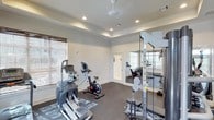 The fitness center at Dozier is a thoughtfully designed workout space that combines functionality with a clean, polished aesthetic. The room features a tray ceiling with recessed lighting, which provides even, bright illumination throughout the space, creating an energizing environment for exercise. Crown molding adds a refined architectural detail that elevates the overall appearance of the room.

The facility is outfitted with a variety of cardio equipment, including an elliptical trainer and a stationary cycle, positioned to allow comfortable use. A multi-station strength training rack with weight plates is situated along one wall, offering users a range of resistance training options. Durable rubber flooring covers the ground, providing both safety and cushioning during workouts.

A large wall mirror extends across one side of the room, visually expanding the space while allowing users to monitor their form. Natural light filters in through windows fitted with blinds, supplementing the recessed overhead lighting. A doorway leads to an adjacent area, suggesting additional fitness or utility space beyond. The neutral white walls and clean layout contribute to an organized, focused atmosphere well-suited for daily exercise.