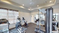 The fitness center at Dozier is a thoughtfully designed workout space that combines functionality with a clean, polished aesthetic. The room features a tray ceiling with recessed lighting, which provides even, bright illumination throughout the space, creating an energizing environment for exercise. Crown molding adds a refined architectural detail that elevates the overall appearance of the room.

The facility is outfitted with a variety of cardio equipment, including an elliptical trainer and a stationary cycle, positioned to allow comfortable use. A multi-station strength training rack with weight plates is situated along one wall, offering users a range of resistance training options. Durable rubber flooring covers the ground, providing both safety and cushioning during workouts.

A large wall mirror extends across one side of the room, visually expanding the space while allowing users to monitor their form. Natural light filters in through windows fitted with blinds, supplementing the recessed overhead lighting. A doorway leads to an adjacent area, suggesting additional fitness or utility space beyond. The neutral white walls and clean layout contribute to an organized, focused atmosphere well-suited for daily exercise.