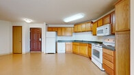 This single-floor residential unit offers a functional and practical layout designed for comfortable everyday living. The kitchen is a standout feature, equipped with warm oak cabinetry providing ample upper and lower storage, dark laminate countertops, a double-basin sink, and white appliances including a range, over-the-range microwave, and dishwasher. The kitchen flows openly into the adjacent living area, which is finished with neutral carpeting and bright white walls, creating a cohesive and airy feel. Overhead fluorescent lighting illuminates the space effectively throughout.

The unit includes a carpeted bedroom with a window fitted with horizontal blinds, allowing for natural light and outside views. The floor plan, visible in the dollhouse view, reveals a well-organized corridor connecting the living spaces to the private areas of the unit. Two bathrooms serve the unit — one features a walk-in shower, an oak vanity with a light countertop, a large mirror, and tile flooring, while the second includes a full bathtub, matching oak cabinetry, and tile flooring. A dedicated in-unit laundry room houses a full-sized side-by-side washer and dryer with wire shelving above. Throughout the unit, consistent use of oak finishes and neutral tones contributes to a cohesive and practical interior aesthetic.