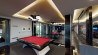 Located at 5225 Las Colinas Blvd in Irving, Texas, this contemporary two-floor hospitality property offers a well-appointed and dynamic environment designed for comfort and social engagement. The grand lobby immediately impresses with soaring double-height ceilings, floor-to-ceiling windows adorned with bold black-and-white striped curtains, and an abundance of natural light. Oversized pendant lighting in deep navy anchors the space, while a stone fireplace flanked by seating areas and wall-mounted displays creates a warm, inviting atmosphere. A communal wood dining table serves as a central gathering point. Adjacent to the lobby, a convenience market provides retail refrigerated displays and snack offerings for guests. A glass-enclosed staircase with polished metal railings connects the two floors, leading to an upper level overlooking the main space. The game lounge is a standout feature, housing a striking red-felted billiards table with a sculptural base, complemented by modern multi-shade pendant lighting, glossy dark tile flooring, and cove-lit tray ceilings. Card and bar-height seating areas, a shuffleboard table, and large windows with outdoor views complete the entertainment zone. Throughout, curated abstract artwork, dark reflective columns, and cohesive contemporary finishes contribute to a polished, upscale aesthetic.