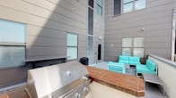 Located at 15 E Lane Ave in Columbus, Ohio's University District, this outdoor courtyard amenity space is thoughtfully designed for social gatherings and relaxation. The courtyard is enclosed by a contemporary multi-story building exterior clad in sleek gray panel siding, which rises impressively overhead and creates a sense of privacy while allowing ample open sky above.

At the heart of the space is a built-in outdoor kitchen area, anchored by a large stainless steel gas grill mounted on a tiled countertop with warm wood-toned accents — ideal for entertaining. Adjacent to the grilling station, a comfortable lounge area is furnished with vibrant teal upholstered chairs and seating, adding a lively pop of color against the neutral architectural backdrop.

The building's facade features multiple windows and a centrally positioned door providing direct access to the interior, making the transition between indoor and outdoor spaces seamless. Security and utility elements are discreetly integrated throughout the design. The overall ambiance of the courtyard is modern and functional, balancing clean architectural lines with welcoming outdoor living amenities — making it a practical and stylish communal retreat for residents.