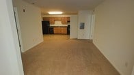 This single-floor unit offers an open-concept layout that seamlessly connects the living area to the kitchen, creating a practical and functional living space. The generously sized living area is finished with neutral wall-to-wall carpeting, providing a clean and understated foundation that suits a variety of furnishing styles.

The kitchen is positioned at the rear of the space and is distinguishable by its tile flooring, which provides a clear transition from the carpeted living zone. It features warm wood-tone cabinetry both above and below the counter, offering ample storage. Black appliances, including a full-size refrigerator, an oven range, and a dishwasher, are integrated throughout. A sink is centrally located along the countertop, and overhead lighting illuminates the workspace effectively.

An electrical panel box is visible on the wall adjacent to the kitchen, and a doorway nearby leads to additional areas of the unit. The overall aesthetic is straightforward and functional, with neutral-toned walls throughout that contribute to a bright and open atmosphere. The layout is well-suited for efficient day-to-day living.