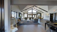 The Timbers presents a thoughtfully designed single-floor space that immediately impresses with its dramatic vaulted ceiling, defined by striking exposed dark beams that draw the eye upward and create a strong architectural identity. The open-plan layout offers a generous, flowing interior that seamlessly connects multiple seating and gathering areas without visual interruption.

A large gable-end window wall at the far end of the space floods the interior with abundant natural light, complemented by additional windows along the side walls that provide views of the surrounding outdoor landscape. French doors near the window wall enhance the connection between indoors and outdoors.

The interior is finished with smooth concrete-style flooring throughout, providing a neutral, contemporary foundation. Furnishings are modern and carefully curated, including a gray sectional sofa, velvet accent chairs in deep green, and a dining area appointed with mustard-toned seating. A sculptural multi-globe chandelier serves as a focal point overhead, adding an artistic touch.

The overall ambiance is warm yet contemporary, blending industrial and modern design elements to create an inviting, versatile communal environment.