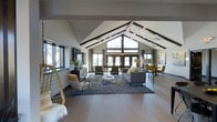 The Timbers presents a thoughtfully designed single-floor space that immediately impresses with its dramatic vaulted ceiling, defined by striking exposed dark beams that draw the eye upward and create a strong architectural identity. The open-plan layout offers a generous, flowing interior that seamlessly connects multiple seating and gathering areas without visual interruption.

A large gable-end window wall at the far end of the space floods the interior with abundant natural light, complemented by additional windows along the side walls that provide views of the surrounding outdoor landscape. French doors near the window wall enhance the connection between indoors and outdoors.

The interior is finished with smooth concrete-style flooring throughout, providing a neutral, contemporary foundation. Furnishings are modern and carefully curated, including a gray sectional sofa, velvet accent chairs in deep green, and a dining area appointed with mustard-toned seating. A sculptural multi-globe chandelier serves as a focal point overhead, adding an artistic touch.

The overall ambiance is warm yet contemporary, blending industrial and modern design elements to create an inviting, versatile communal environment.