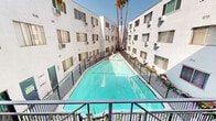 Located in the heart of Hollywood at 1811 Tamarind Ave, this single-floor residence is part of a multi-story complex designed around a striking central courtyard. The standout feature of the property is the expansive, resort-style swimming pool that stretches the length of the courtyard, enclosed by a black iron fence and framed by soaring palm trees against the classic Southern California sky. The white stucco exterior gives the complex a clean, bright aesthetic consistent with mid-century Los Angeles architecture.

The unit includes a generously sized private balcony that overlooks the pool and courtyard below, offering an inviting outdoor living area. The balcony is outfitted with wooden lounge seating, making it an ideal spot for relaxing while enjoying views of the lush palm tree canopy. A set of full-height glass sliding panels connects the balcony to the interior, allowing abundant natural light to fill the space and creating a seamless indoor-outdoor flow.

The covered balcony corridor also provides access to unit entryways, adding a semi-private transitional space between the shared amenities and individual residences. The overall ambiance of the property blends practical urban living with leisure-oriented outdoor amenities, well-suited to the vibrant Hollywood neighborhood it calls home.