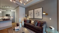 This thoughtfully designed single-floor residence combines contemporary style with comfortable living across a well-appointed layout. The open-concept living area is a standout, anchored by a tray ceiling with elegant crown molding and an eye-catching branching chandelier with globe bulbs. Warm-toned hardwood flooring runs throughout the main living spaces, complementing the deep charcoal walls that lend a sophisticated, moody ambiance. A large sectional sofa, paired abstract framed artwork, and brass wall sconces complete the inviting atmosphere.

The kitchen features sage-gray cabinetry, granite countertops, a classic subway tile backsplash, and stainless steel appliances including a French door refrigerator. A breakfast bar with industrial-style seating provides a casual dining option. The entry foyer is clean and functional, with a security panel and direct sightlines into the kitchen.

The bedroom offers a tranquil retreat, dressed in soft, layered neutral tones with a tufted upholstered headboard and bold accent artwork. Floor-to-ceiling windows frame lush outdoor tree views, filling the room with natural light. A hallway with mirrored closet doors leads to a well-designed bathroom featuring blue-gray cabinetry and an arched architectural detail, adding a refined touch to the private quarters.