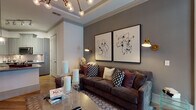 This thoughtfully designed single-floor residence combines contemporary style with comfortable living across a well-appointed layout. The open-concept living area is a standout, anchored by a tray ceiling with elegant crown molding and an eye-catching branching chandelier with globe bulbs. Warm-toned hardwood flooring runs throughout the main living spaces, complementing the deep charcoal walls that lend a sophisticated, moody ambiance. A large sectional sofa, paired abstract framed artwork, and brass wall sconces complete the inviting atmosphere.

The kitchen features sage-gray cabinetry, granite countertops, a classic subway tile backsplash, and stainless steel appliances including a French door refrigerator. A breakfast bar with industrial-style seating provides a casual dining option. The entry foyer is clean and functional, with a security panel and direct sightlines into the kitchen.

The bedroom offers a tranquil retreat, dressed in soft, layered neutral tones with a tufted upholstered headboard and bold accent artwork. Floor-to-ceiling windows frame lush outdoor tree views, filling the room with natural light. A hallway with mirrored closet doors leads to a well-designed bathroom featuring blue-gray cabinetry and an arched architectural detail, adding a refined touch to the private quarters.