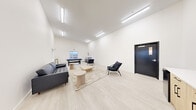 This contemporary lounge and recreation space is thoughtfully designed to offer both comfort and entertainment in a clean, minimalist aesthetic. The single-floor layout is defined by impressively high ceilings that amplify the sense of openness, while bright white walls and recessed linear lighting create a crisp, well-lit atmosphere throughout.

The main living area serves as the social heart of the space, featuring light wood-style flooring that adds warmth to the modern design. A tufted black leather sofa anchors a comfortable seating arrangement, complemented by sleek black accent chairs and a combination of low-profile natural wood coffee and side tables arranged atop a neutral-toned area rug. A pool table positioned near a window at the far end of the room introduces a recreational element, with natural light and exterior views providing an inviting backdrop.

A small built-in cabinetry unit and countertop area offer practical storage and utility. The space also includes an accessible bathroom, finished with neutral stone-look tile flooring, a wall-mounted sink with a light wood vanity, a framed mirror, and standard fixtures — all maintaining the clean, functional design language of the overall space. A dark-framed entry door adds a bold contrast to the predominantly white interior, completing the modern look.