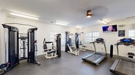 Located at 6001 Thomaston Rd in Macon, Georgia, this fitness center offers a well-equipped, single-floor exercise space designed to support a full range of workout routines. The room features clean white walls accented with crown molding, durable rubber flooring, and ample overhead fluorescent lighting supplemented by a ceiling fan, creating a bright and comfortable environment for physical activity.

The cardio zone is equipped with multiple treadmills and an elliptical trainer, all positioned near windows with blinds that allow natural light to filter in from multiple sides of the room. Wall-mounted televisions provide entertainment during workouts. A large mirror spans one wall, visually expanding the space and allowing users to monitor their form.

The strength training area includes several cable and weight machines suited for a variety of muscle groups, along with a dedicated free weights section featuring a tiered dumbbell rack and a flat workout bench. Elegant French doors with glass pane inserts serve as an entry point, adding a refined architectural touch to the otherwise utilitarian fitness space.

Overall, the exercise room is thoughtfully organized to accommodate both cardio and strength training needs, offering residents a convenient and fully functional facility for their daily fitness routines.