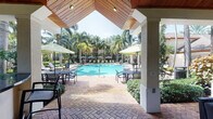 This single-story outdoor pavilion is designed as a fully appointed resort-style amenity space, combining thoughtful architectural detail with a tropical ambiance. The structure is anchored by a striking vaulted ceiling clad in warm natural wood, supported by painted stucco columns that frame open views of the surrounding landscape on all sides.

The pavilion is divided into two primary functional zones. The first is a curved outdoor bar with a dark granite countertop, pendant lighting, and barstool seating — positioned to overlook the expansive pool deck. Adjacent to the bar, a grilling station adds full outdoor kitchen functionality. The second zone serves as an open-air lounge, featuring a freestanding fireplace with a stainless steel surround, a wall-mounted flat-screen television, ceiling fans, and comfortable wicker seating with cushions.

Throughout the space, herringbone-patterned brick pavers provide a cohesive and durable flooring surface. The surrounding pool deck offers multiple seating areas shaded by large market umbrellas, all set against a backdrop of tall palm trees and manicured tropical plantings. The open-air design ensures abundant natural light and cross-ventilation, creating a comfortable and inviting gathering environment year-round.