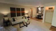 This single-floor residence showcases an open-concept design that seamlessly connects the living and dining areas, creating a cohesive and functional layout. The living area features plush wall-to-wall carpeting underfoot, providing a soft contrast to the hardwood flooring that extends into the dining space beyond. A neutral color palette of warm beiges and taupes forms the backdrop, complemented by a bold charcoal accent wall in the dining area that adds depth and visual interest.

The living room is anchored by a contemporary sofa paired with a sleek glass-top coffee table with dark wood framing. A striking triptych artwork in shades of brown, orange, and black adorns the wall, adding a bold artistic focal point. Soft ambient lighting from a table lamp enhances the room's warm atmosphere.

The adjoining dining area features a round dark wood table with upholstered chairs, lit by an elegant ceiling fan with integrated lighting. Decorative mirrors and floral arrangements on the accent wall contribute to a polished, curated aesthetic. A hallway visible in the background hints at additional rooms within the residence, completing this well-designed living environment.