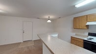 This single-floor space offers a clean, open layout designed for functional everyday living. The kitchen serves as a central highlight, anchored by a generously sized island with speckled stone-style countertops that extend into an eat-in or workspace overhang. A classic pendant light hangs above, providing focused illumination and adding a subtle design accent to the area. Warm-toned wood cabinetry lines the surrounding walls, offering ample storage while complementing the light, neutral color scheme throughout. The flooring transitions from tile in the kitchen zone to carpet in the adjacent living area, clearly defining each functional space. Walls are painted in a crisp white throughout, contributing to a bright and airy ambiance. A solid paneled entry door with standard hardware provides access from the exterior, and recessed ceiling vents suggest a central HVAC system for year-round climate control. The overall design is straightforward and practical, with a consistent neutral palette that provides a versatile backdrop suitable for a variety of furnishing styles. The open flow between the kitchen and living areas creates a connected, cohesive feel across the single level.