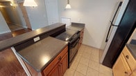 This single-floor residence is thoughtfully designed with a functional layout that connects living, cooking, and sleeping areas seamlessly. The space features two distinct kitchen zones: one with warm-toned medium wood cabinetry paired with dark granite-style countertops, an electric range, and pendant lighting, and a second galley-style kitchen with light maple cabinetry, dark laminate countertops, stainless steel sink, black refrigerator, and built-in microwave. Both kitchens are finished with neutral ceramic tile flooring.

The living area showcases rich hardwood flooring that extends into the bedrooms, complemented by neutral wall tones and large windows that invite generous natural light. A set of French-style doors opens to a balcony, further enhancing the bright and open feel. The bedroom features dual windows with views of the surrounding neighborhood and ample wall space.

Two bathrooms serve the unit: one features a full tub-shower combination, a pedestal sink with chrome fixtures, a vanity mirror with elegant lighting, and a window for natural ventilation. The second bathroom offers a walk-in tiled shower stall with light ceramic tile flooring, providing a clean and practical design.
