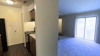 This single-floor residential unit offers a straightforward and functional layout designed for comfortable everyday living. The main living area is finished with plush wall-to-wall carpet in a neutral tone, providing a warm and inviting atmosphere. Large windows allow natural light to fill the space, enhancing the open feel of the room. Smooth, light-gray walls and clean white baseboards contribute to the unit's fresh, move-in-ready appearance.

The kitchen is positioned adjacent to the living area, separated by a structural column that defines each zone while maintaining a sense of connectivity. The kitchen features dark wood cabinetry, a built-in electric range with an overhead range hood, and dark hardwood-style flooring that contrasts with the carpeted living space. The cabinetry provides ample storage, and the layout supports efficient use of the cooking area.

A hallway visible from the living room leads to additional rooms beyond, suggesting a multi-room floor plan suited for various living arrangements. Ceiling vents indicate a central HVAC system throughout the unit. Overall, the space presents a clean, neutral palette with practical finishes, making it a versatile and functional living environment.
