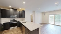 Located in Skillman, NJ, this well-appointed single-floor residence offers three bedrooms, two bathrooms, and a smartly designed layout that maximizes functionality and comfort. The modern kitchen is a standout feature, showcasing dark shaker-style cabinetry, granite-look countertops, a central island with a built-in sink, and a full suite of stainless steel appliances including a gas range, microwave, and refrigerator with ice maker. Light wood-look flooring flows throughout the main living areas, complementing the clean white walls and recessed lighting overhead. The open-concept design connects the kitchen seamlessly to the living room, which features sliding glass doors leading to a private balcony. The spacious primary bedroom includes a walk-in closet with built-in wire shelving, while additional bedrooms offer plush carpeting and ample natural light. Both bathrooms are finished with dark vanity cabinetry, white countertops, and tile flooring; one features a tub-shower combination with white subway tile surround. A dedicated utility room and hallway complete the layout. The overall aesthetic is contemporary and move-in ready, with neutral tones and quality finishes throughout.