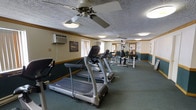 This fitness room offers a functional and well-organized workout space designed to accommodate a variety of exercise needs. The room is generously sized, providing ample space for multiple users to exercise comfortably without feeling cramped. The flooring is covered in teal-toned carpet, lending the space a clean, defined aesthetic. A textured ceiling with multiple ceiling fans ensures consistent air circulation throughout the area, contributing to a comfortable workout environment.

The cardio section features a row of treadmills, including a prominent Horizon T80 model, lined along one side of the room. Toward the rear of the space, a multi-station strength training machine provides options for resistance and weight-based exercises. Wall-mounted air conditioning units support climate control, supplementing the ceiling fans for year-round comfort.

Natural light filters in through windows dressed with vertical blinds, brightening the space without creating glare. Decorative crown molding and framed artwork add a subtle sense of polish to what is otherwise a practical, utility-focused room. Overall, the fitness room strikes a balance between functionality and a welcoming atmosphere suitable for daily use.
