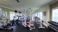 This fitness center is designed to support a full range of exercise routines in a clean, organized environment. The space is outfitted with a multi-station cable machine at its center, complemented by a variety of strength-training equipment featuring distinctive orange-padded seats and benches. Cardio machines, including treadmills and a stationary bike, are positioned along one side of the room, offering users a well-rounded workout experience. The rubber flooring throughout provides both durability and safety underfoot. Large windows along the perimeter flood the space with natural light, creating an open and energizing atmosphere, while slatted blinds offer adjustable light control. Full-length mirrors lining the walls enhance the sense of spaciousness and allow users to monitor their form during workouts. The white walls and light-colored ceiling contribute to a clean, airy aesthetic. Motivational fitness imagery is displayed on the walls, adding to the purpose-driven ambiance. A ceiling fan provides additional air circulation, ensuring comfort during intense workouts. Overall, the space is thoughtfully arranged to maximize usability while maintaining an uncluttered, welcoming environment suited for a wide range of fitness activities.