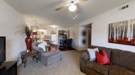 This single-floor residence offers a welcoming open-concept layout that seamlessly connects the main living, dining, and kitchen areas into one cohesive space. The living area is generously sized, featuring plush wall-to-wall carpeting that adds warmth underfoot. A ceiling fan with a light fixture is centrally mounted, providing both airflow and overhead illumination, while a textured ceiling adds a classic finishing touch. The space accommodates comfortable seating arrangements, making it ideal for relaxing or entertaining. Adjacent to the living area, the dining space offers room for a full dining set, creating a defined yet open area for meals. The kitchen is visible from both the living and dining zones, featuring light cabinetry, a full-size refrigerator, and ample counter space, reinforcing the connected, functional nature of the floor plan. A hallway leads to additional rooms beyond, offering privacy from the main living areas. The neutral wall tones throughout create a bright, clean backdrop that complements a variety of décor styles. Decorative wall accents and thoughtful furnishing arrangements highlight the versatility and livability of this single-level home.