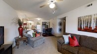 This single-floor residence offers a welcoming open-concept layout that seamlessly connects the main living, dining, and kitchen areas into one cohesive space. The living area is generously sized, featuring plush wall-to-wall carpeting that adds warmth underfoot. A ceiling fan with a light fixture is centrally mounted, providing both airflow and overhead illumination, while a textured ceiling adds a classic finishing touch. The space accommodates comfortable seating arrangements, making it ideal for relaxing or entertaining. Adjacent to the living area, the dining space offers room for a full dining set, creating a defined yet open area for meals. The kitchen is visible from both the living and dining zones, featuring light cabinetry, a full-size refrigerator, and ample counter space, reinforcing the connected, functional nature of the floor plan. A hallway leads to additional rooms beyond, offering privacy from the main living areas. The neutral wall tones throughout create a bright, clean backdrop that complements a variety of décor styles. Decorative wall accents and thoughtful furnishing arrangements highlight the versatility and livability of this single-level home.