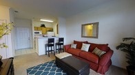 This single-floor space features an open-concept design that seamlessly connects the living area and kitchen. The living room is highlighted by plush carpeting, a rich red sofa accented with decorative pillows, and a dark leather ottoman resting atop a teal geometric-patterned area rug. A framed mirror with a rustic wood border adorns the neutral-toned wall, adding depth to the room.

The kitchen is positioned adjacent to the living area and is separated by a peninsula breakfast bar equipped with two black bar stools, creating a natural divide while maintaining an open feel. The kitchen features warm honey-toned wood cabinetry, white appliances including a microwave and refrigerator, and light-colored countertops. A hanging fluorescent light fixture illuminates the kitchen workspace. Tile flooring in the kitchen transitions to carpeting in the living space. The overall ambiance is warm and functional, with a practical layout that maximizes the use of the available space, making it well-suited for everyday living.