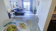 This well-appointed single-floor residence offers an open-concept layout that seamlessly connects the dining and living areas, creating a spacious and inviting atmosphere. Wood-style plank flooring runs throughout, lending a warm yet contemporary aesthetic to the entire space.

The living area is bathed in natural light streaming through large sliding glass doors that lead to a private balcony, offering views of the surrounding greenery. The space is generously proportioned, easily accommodating a full sectional sofa, coffee table, and additional seating. Soft, flowing curtains frame the glass doors, adding an elegant touch to the room.

The dining area sits just adjacent to the living space, centered around a round glass-top table with modern seating — a sleek arrangement that maintains an open, airy feel without compromising functionality.

Throughout the space, neutral gray walls serve as a versatile backdrop, complemented by tasteful pops of color in the décor, including blue accent pillows and vibrant artwork. The overall ambiance is bright, clean, and contemporary, with thoughtful design choices that maximize both comfort and style. The private balcony extends the living space outdoors, perfect for enjoying fresh air and natural surroundings.