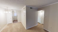 Located at 9371 Mansfield Rd in Shreveport, Louisiana, this single-floor unit offers a practical and comfortable layout throughout. The living area is generously proportioned, featuring plush carpet flooring, crown molding, and a large front-facing window that allows natural light to fill the space. A standout feature is the white-painted exposed brick accent wall, adding a touch of classic character to the interior. An elegant chandelier fixture highlights the dining area, which connects seamlessly to the rest of the unit via a well-defined layout visible in the dollhouse view. The galley-style kitchen is efficiently designed with white cabinetry, laminate countertops, a double-basin stainless steel sink, white appliances including a coil-top range and refrigerator, and an over-range ventilation hood. A window above the sink provides natural light to the kitchen. Hardwood-style flooring runs through the kitchen and transition areas, offering a clean contrast to the carpeted living spaces. The bathroom features a vanity with a large mirror, scalloped light fixture, and an adjacent tub-shower combination with tile surround. Textured ceilings with crown molding details are consistent throughout the unit, contributing to a cohesive and tidy aesthetic.