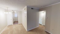 Located at 9371 Mansfield Rd in Shreveport, Louisiana, this single-floor unit offers a practical and comfortable layout throughout. The living area is generously proportioned, featuring plush carpet flooring, crown molding, and a large front-facing window that allows natural light to fill the space. A standout feature is the white-painted exposed brick accent wall, adding a touch of classic character to the interior. An elegant chandelier fixture highlights the dining area, which connects seamlessly to the rest of the unit via a well-defined layout visible in the dollhouse view. The galley-style kitchen is efficiently designed with white cabinetry, laminate countertops, a double-basin stainless steel sink, white appliances including a coil-top range and refrigerator, and an over-range ventilation hood. A window above the sink provides natural light to the kitchen. Hardwood-style flooring runs through the kitchen and transition areas, offering a clean contrast to the carpeted living spaces. The bathroom features a vanity with a large mirror, scalloped light fixture, and an adjacent tub-shower combination with tile surround. Textured ceilings with crown molding details are consistent throughout the unit, contributing to a cohesive and tidy aesthetic.