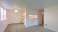 This single-floor residence offers a clean, open layout designed for comfortable everyday living. The living area is finished with neutral carpeting that adds warmth underfoot, while a ceiling fan with integrated lighting provides both air circulation and ambient illumination. A partial dividing wall separates the living space from the kitchen, incorporating a pass-through window that maintains visual connectivity between the two areas and allows for easy communication and serving. The kitchen features light wood cabinetry, tile flooring, and ample counter space, giving it a practical and functional character. The cabinetry extends to the upper walls, offering generous storage. The overall color palette throughout the space is light and neutral, with white walls that enhance the sense of openness and allow natural light to reflect freely. The single-floor plan ensures easy accessibility and a straightforward flow between spaces. The interior presents a well-maintained, move-in ready environment with a simple, understated aesthetic that provides a versatile backdrop for a variety of furnishing styles.