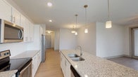 This well-appointed single-story residence combines modern finishes with a thoughtfully designed open layout. The kitchen serves as the centerpiece of the home, showcasing a large central island with granite countertops, a deep double-basin sink, and elegant pendant lighting overhead. White shaker-style cabinetry lines the walls, complemented by a subway tile backsplash and a full suite of stainless steel appliances, including a range, microwave, and refrigerator. Light wood-style flooring flows seamlessly through the kitchen and into the open living and dining areas, where a ceiling fan and glass doors invite natural light and outdoor access. The bedroom features plush carpeting, a ceiling fan, and wide windows with outdoor views, creating a calm and comfortable retreat. The primary bathroom is generously sized, offering a dual-sink vanity with granite countertops, a glass-enclosed shower, and light wood-tone flooring. A secondary bathroom provides a single-sink granite vanity with coordinating finishes. An in-unit laundry area rounds out the functional amenities of this home. Throughout the space, a neutral color palette and clean architectural lines create a bright, cohesive, and move-in-ready environment.