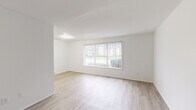 This single-floor unit features a clean, minimalist design with a bright, white interior palette throughout. The open living area is finished with light-toned wood laminate flooring and anchored by a large picture window fitted with vertical blinds, allowing abundant natural light and views of the surrounding greenery. A secondary living or dining area shares the same flooring and benefits from an additional large window with vertical blinds along one wall, further enhancing the airy atmosphere.

The galley-style kitchen is efficiently laid out with white cabinetry on both sides, light quartz-style countertops, and a stainless steel sink. Appliances include a black and stainless electric range with overhead ventilation and a stainless refrigerator, providing a functional cooking environment. Light wood laminate flooring continues here, maintaining visual consistency.

The bedrooms feature neutral carpet flooring for added comfort. One bedroom is well-lit by two windows with horizontal blinds positioned on adjacent walls, while another includes a wall-mounted ductless mini-split unit for individual climate control. White walls throughout the unit contribute to a clean, move-in-ready presentation. A bathroom is also present, as indicated in the layout overview.