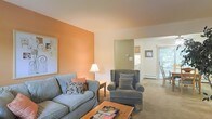 This single-floor space centers around a comfortable living area that immediately draws attention with its bold terracotta-orange accent wall, which adds warmth and character against the neutral cream ceiling and surrounding walls. The living room is furnished with a classic sofa and a matching wingback armchair, both upholstered in soft blue-grey fabric, creating a relaxed and cohesive seating arrangement. A rustic wooden coffee table anchors the space, while a traditional floor lamp provides ambient lighting. Neutral-toned carpeting runs throughout, contributing to the overall cozy atmosphere. An open passageway leads from the living area into what appears to be a kitchen, distinguished by its sage-green accent wall and glimpses of cabinetry. A second opening connects to a brighter room featuring a window with natural light filtering through, suggesting a dining area or additional living space. Framed artwork adorns the walls, adding a personal touch. The layout flows naturally between spaces, offering a functional and welcoming environment suited for everyday living.