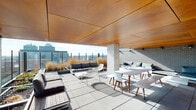 This rooftop terrace is a well-designed outdoor amenity space that seamlessly blends covered and open-air areas, offering a versatile environment for socializing and relaxation. The architecture is defined by a striking warm-toned wood panel ceiling that extends over a substantial portion of the terrace, providing shade while adding a refined, modern aesthetic. Exposed concrete columns support the overhead structure, contributing an industrial character that complements the white brick accent walls found throughout the space.

The terrace is organized into distinct functional zones. A lounge area features a generously sized sectional sofa with deep cushions, positioned against a white brick wall beneath the wood ceiling, with a wall-mounted flat-screen TV and track lighting overhead — creating an inviting outdoor media and gathering spot. Raised planter beds filled with ornamental grasses introduce natural texture and greenery, acting as design dividers within the space.

A dining zone accommodates multiple guests with long white tables and modern wood-legged chairs, bathed in natural light from the open sky above. Large-format stone pavers cover the floor throughout, providing a clean and durable surface. The terrace opens to unobstructed panoramic views of the surrounding cityscape and skyline, flooding the space with natural light and enhancing the elevated, airy ambiance of this thoughtfully appointed outdoor retreat.