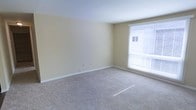 This single-floor space features a well-proportioned room finished with soft, neutral cream-colored walls and clean white baseboards that create a bright, understated aesthetic. The room is carpeted in a light beige tone, adding warmth and comfort underfoot throughout the space. A window fitted with horizontal blinds allows natural light to filter in, illuminating the room and offering a view of the surrounding exterior. The walls include standard electrical outlets, reflecting a practical, functional layout. The overall ambiance is calm and move-in ready, providing a blank canvas well-suited for a variety of furnishing arrangements and personal styles.