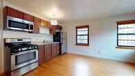 This single-floor unit offers a thoughtfully designed layout that combines functionality with classic charm. Warm hardwood flooring runs continuously throughout the space, creating a cohesive and inviting atmosphere from room to room.

The kitchen is a standout feature, outfitted with rich medium-toned wood cabinetry extending to the ceiling for ample storage, granite-style countertops, and a full suite of stainless steel appliances including a gas range, over-the-range microwave, and refrigerator. A decorative drum pendant light fixture adds a stylish focal point above. The kitchen opens naturally into the living area, forming a practical and airy open-concept space. A double-hung window with wood trim brings in natural light and views of the surrounding exterior.

The bedroom is generously proportioned, featuring corner placement of two windows that allow for abundant natural light. A cast-iron radiator beneath the windows provides efficient radiant heating, complementing the room's warm character.

The bathroom is cleanly finished with white subway tile walls accented by a decorative mosaic tile border in earthy tones. It includes a full soaking tub with overhead shower, a pedestal-style vanity, and a window that allows for natural ventilation. The overall design of the space balances classic building character with modern, updated finishes throughout.
