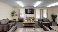 Located at 6501 Hil Mar Dr in District Heights, Maryland, this single-floor community space is thoughtfully designed to provide a welcoming and functional environment. The layout flows naturally from one area to the next, offering a variety of spaces suited for relaxation, socializing, and activity.

The main lounge serves as the heart of the space, featuring plush leather seating arranged around a rustic wood coffee table, a wall-mounted flat-screen television, and multiple windows dressed with patterned drapes that invite natural light. An adjacent sitting nook offers a quieter retreat, furnished with upholstered armchairs, a bookshelf stocked with games and reading materials, and warm ambient lighting.

The dining area accommodates small groups at bistro-style round tables with cushioned bar chairs, accented by contemporary wall art and natural light from surrounding windows. The kitchenette is equipped with dark espresso cabinetry, granite-style countertops, a stainless steel sink, and a full-size refrigerator, all set atop warm hardwood-style flooring.

A spacious multipurpose room provides an open, carpeted area with multiple closets and direct exterior access, offering flexibility for various programming needs. The accessible bathroom is generously sized, featuring tile flooring, grab bars, a wall-mounted sink, and ample vanity space, designed to accommodate all users comfortably. Throughout the space, neutral warm tones and consistent carpeting create a cohesive, inviting atmosphere.