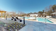 Located at 900 Mountaintop Ave in Gainesville, Georgia, this resort-style outdoor amenity space offers a striking and well-appointed pool environment designed for relaxation and leisure. The centerpiece is a generously sized, crystal-clear swimming pool with a classic rectangular form, complemented by a separate wading or spa area, all finished with polished stone pool decking that extends comfortably around the perimeter.

The pool deck is thoughtfully furnished with an abundance of chaise lounges in both classic black-frame and white styles, arranged in orderly rows and shaded by bold black-and-white striped umbrellas that lend a sophisticated, resort-caliber aesthetic. Distinctive rattan pod lounge chairs add an elegant and stylish touch, providing unique seating options along the poolside.

A handsome stone and brick clubhouse structure anchors one end of the space, featuring arched architectural details, large French doors, and a covered pergola dining area — ideal for gatherings and entertaining. A separate white auxiliary building with private changing areas or restroom facilities is positioned at the opposite end of the pool.

The space is bordered by mature trees and natural Georgia woodland, creating a private, serene atmosphere open to the blue sky above. Manicured flower beds with seasonal blooms add colorful finishing touches, completing this beautifully designed outdoor retreat.