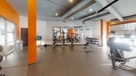Located at 243 Chicago Ave S in Minneapolis, MN, this fitness center is a comprehensively outfitted facility designed to support a wide range of workout routines. The space is organized across a single floor and divided into distinct functional zones, each thoughtfully equipped for different training disciplines.

The main strength training area features rubber flooring, large wall-to-wall mirrors that amplify the sense of space, and a robust selection of equipment including cable machines, a multi-function strength station, adjustable benches, and multiple dumbbell racks. Bold orange accent walls and columns punctuate the otherwise neutral white and gray palette, lending the space an energetic, modern atmosphere. Exposed ductwork runs along the ceiling, contributing to an industrial aesthetic while recessed lighting ensures the area is well lit throughout.

The cardio zone is lined with large windows that allow natural light to pour in, and includes a row of treadmills, stationary bikes, and stair climbers, all equipped with individual monitors.

A separate studio room features light hardwood flooring, floor-to-ceiling mirrors, and a clean, open layout ideal for stretching, yoga, or cycling. An adjoining room with rubber flooring provides space for functional training and houses kettlebells, medicine balls, and stability balls. A wall-mounted flat-screen television adds a modern convenience to the training environment.