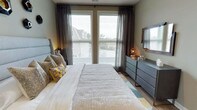 This single-floor residence offers a thoughtfully designed living environment with a contemporary aesthetic that balances comfort and style. The bedroom serves as a standout feature, showcasing a refined interior with large, floor-to-ceiling windows dressed in sheer charcoal curtains and horizontal blinds that filter natural light while framing outdoor views of the surrounding neighborhood. The room is anchored by a generously sized bed with an upholstered gray headboard, layered with textured pillows in warm gold and earthy tones that add a pop of color against the neutral palette. A sleek, dark-toned dresser with gold-finish hardware lines one wall, topped with a mirror and complementary accent pieces that enhance the room's polished look. Warm-toned wall sconces provide ambient lighting, contributing to the inviting atmosphere. The flooring features wood-look planks accented by a geometric area rug in muted tones of teal and gray, tying the room's color story together. The overall ambiance is calm and sophisticated, with a careful attention to detail in both furnishings and finishes that creates a welcoming and visually cohesive space.