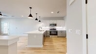 Located at 9520 Whippet Way, Unit 209, in Charlotte, NC, this two-story residence spans 650 square feet, with the primary living space occupying 580 square feet on the upper floor and a 70-square-foot entry level below connected by a staircase. The open-concept kitchen and living area serves as the centerpiece of the unit, showcasing light gray shaker-style cabinetry, quartz countertops, a white subway tile backsplash, and a full suite of stainless steel appliances including a refrigerator, electric range, over-the-range microwave, and dishwasher. A peninsula with an undermount sink and matte black faucet provides additional prep space and casual dining potential. Black pendant lights and recessed ceiling fixtures illuminate the kitchen, while a ceiling fan services the adjacent living area. Light hardwood-style flooring flows throughout the unit, complementing the neutral wall tones and white trim. The bathroom features a floating vanity with drawers, matte black hardware, and matching light wood flooring, maintaining a cohesive modern aesthetic. The dedicated closet and additional doorways visible on the upper floor offer practical storage. Large windows with plantation-style blinds bring in natural light and outdoor views.